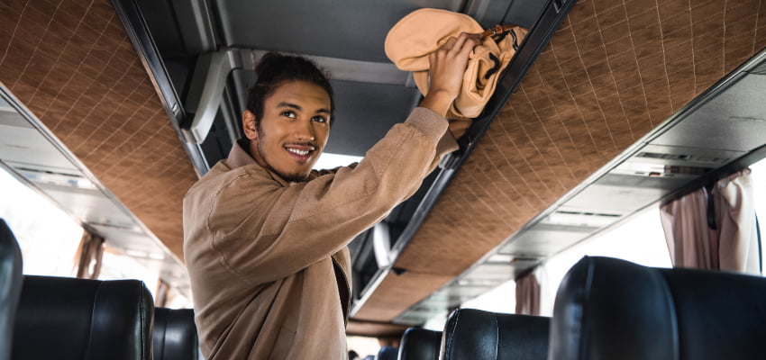 a young man stores his backpack in a charter bus overhead storage bin