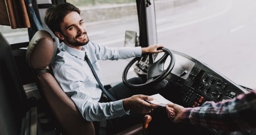 A charter bus driver sits in the driver seat of a bus as a passenger hands them an envelope of money